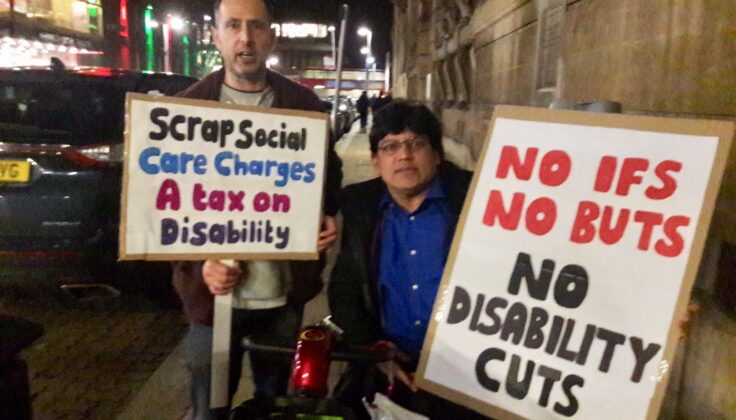 Two individuals are participating in a protest at night outside Redbridge Town Hall. The person on the left is holding a sign that reads "Scrap Social Care Charges A Tax on Disability," and the person on the right is holding a sign saying "NO IFS NO BUTS NO DISABILITY CUTS."