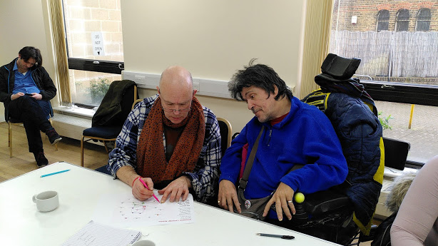 The image captures two individuals engaged in an activity at a table, with one person appearing to assist the other in planning. The individual being assisted is in a blue jacket, seated in a wheelchair,
