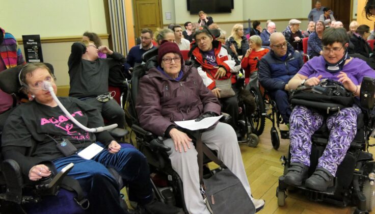 The image depicts a group of campaigners gathered inside Woolwich Town Hall. They are seated, with many individuals using wheelchairs. The group appears to be engaged in an event or a meeting, and the atmosphere suggests they are advocates for a cause.