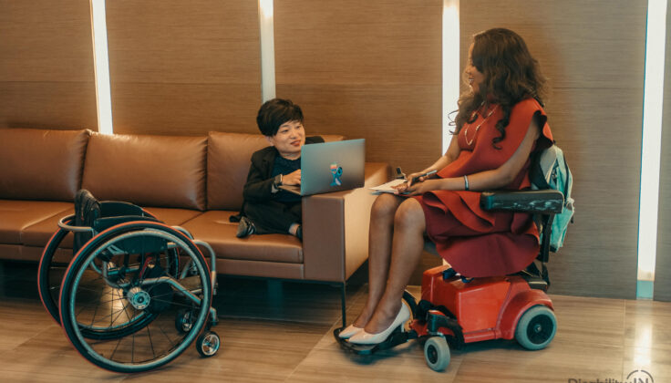 Two women (one Asian woman of restricted height and one Black woman in a motorised scooter) are talking at a sofa