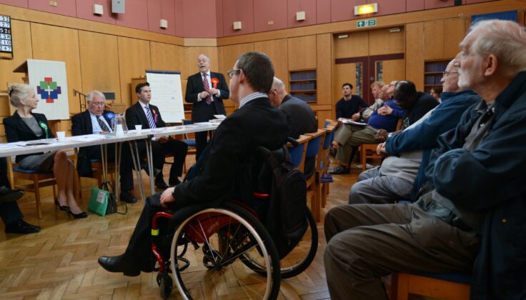 In a well-lit community hall, a diverse audience attentively listens to a panel of political candidates at an election hustings event. A participant in a wheelchair, is in the foreground.