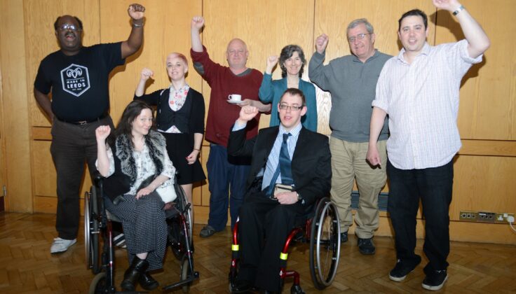 A diverse group of individuals, including some using wheelchairs, are gathered in a room with wood paneling, joyfully raising their fists in a show of unity and empowerment