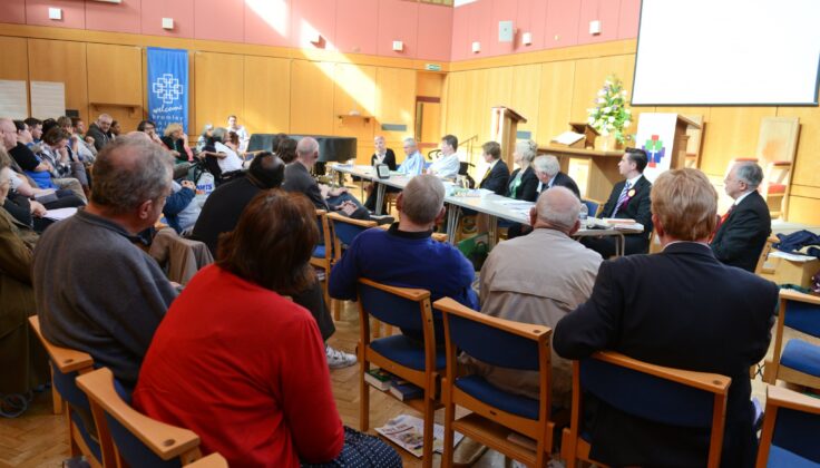 This image shows an election hustings event in an indoor setting. Members of the community are seated in rows, attentively facing a panel of candidates or representatives from different political parties. The candidates are seated behind a long table at the front, likely fielding questions or discussing their platforms. The decor includes wood paneling and acoustic ceiling tiles, indicative of a multi-purpose hall. A projector screen is visible in the background, which may be used to display information relevant to the discussion. Banners on the side suggest the involvement of specific organizations or advocacy groups.