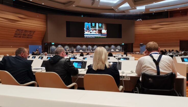 photo from August 2023 evidence session at the UN. a big conference room with dozens of people attending and listening, with a screen at the front showing the speaker and live captions.